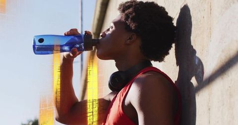 Young athlete drinking water while resting against concrete wall wearing headphones