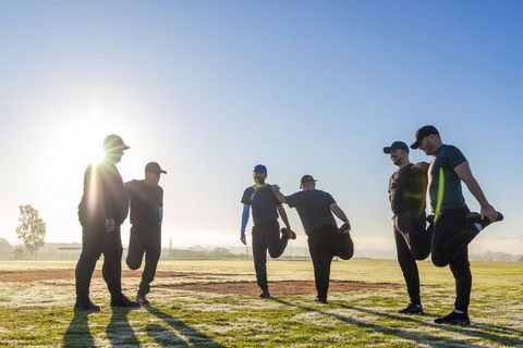 Male teammates stretching on dewy baseball field at sunrise, warming up together