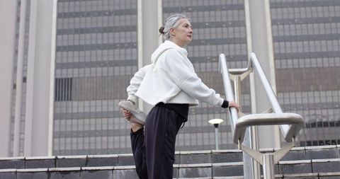 Mature woman stretching quads on urban steps while holding handrail during outdoor workout