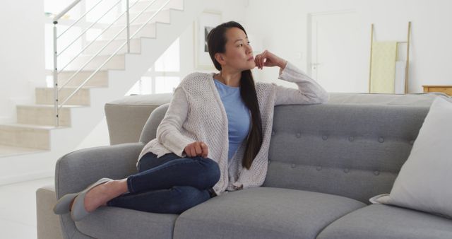 Young woman with long hair sitting on a comfortable gray couch in a modern and minimalistic living room. She is wearing casual wear and looks pensive, resting her chin on her hand. This can be used for themes related to home comfort, relaxation, interior decoration, or reflecting on thoughts. Ideal for articles or commercials about mental well-being, modern living, and lifestyle content.