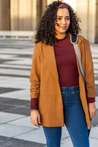 Young woman standing in urban plaza wearing mustard blazer and carrying shoulder bag
