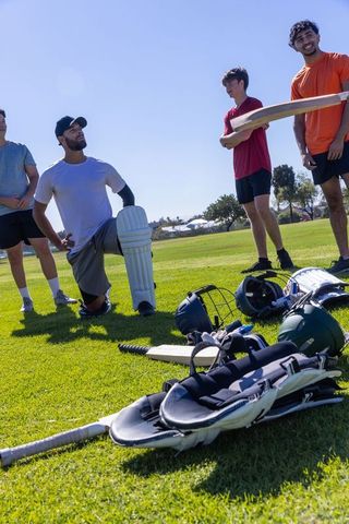 Young men preparing for cricket practice with bats and pads on sunny grass field