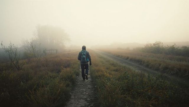 Solo hiker walking misty meadow trail with backpack and beanie at sunrise foggy path