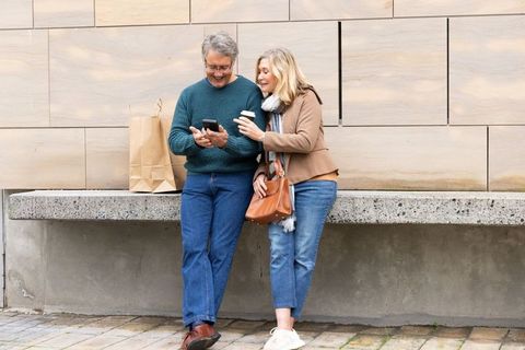 Senior couple leaning on urban bench, smiling and checking smartphone while holding coffee