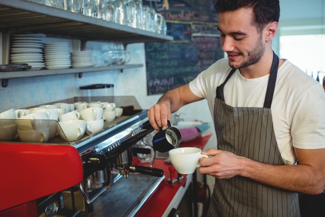 Young male barista pouring coffee into a cup at a cafe. He is wearing an apron and smiling while using an espresso machine. Ideal for content related to coffee shops, barista training, hospitality industry, and small business promotions.