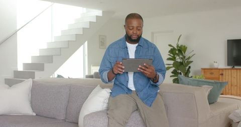 African american man sitting on sofa using tablet wearing denim shirt in modern minimal living room