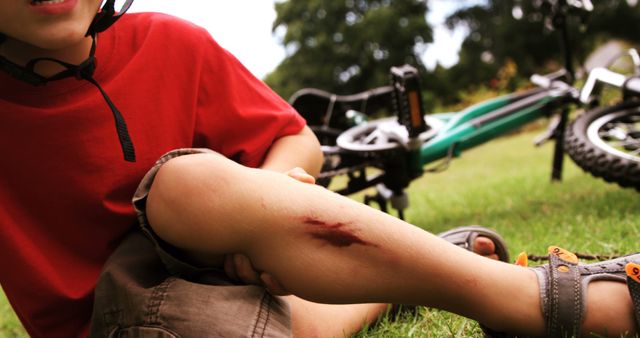 A young Caucasian boy is sitting on the grass showing a scraped knee, with bicycles lying in the background, with copy space. It suggests an outdoor playtime that resulted in a minor injury, emphasizing the importance of safety during physical activities.