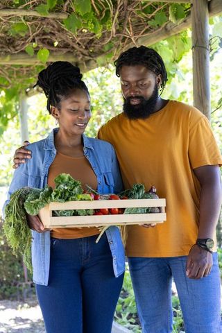 African couple holding crate of fresh homegrown vegetables under sunlit pergola harvesting