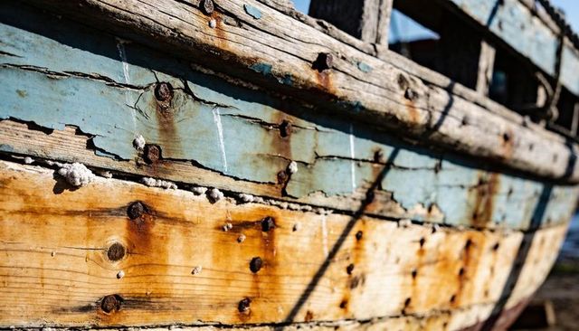Peeling blue paint revealing weathered sunlit boat hull with rusted nails and barnacles