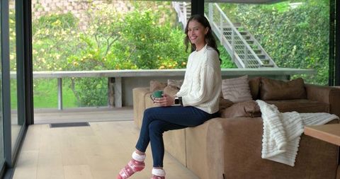 Cozy morning at home woman smiling on sofa arm sipping coffee in knit sweater and festive socks