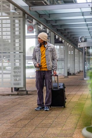 Young traveler standing with suitcase and phone under glass canopy at urban transit terminal wearing