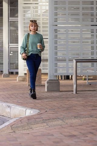 Woman walking with coffee across brick plaza by modern urban architecture, casual style