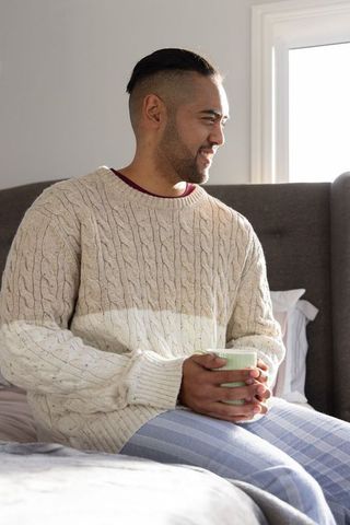 Mid-adult asian man enjoying quiet morning holding mug on bed in cozy cable-knit sweater