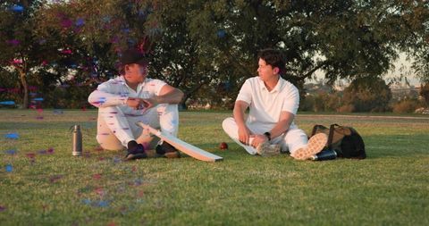 Two cricket teammates sitting on park grass chatting during break with bat and red ball