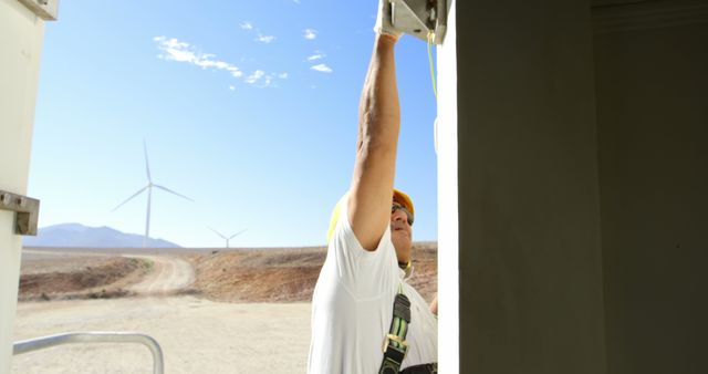 Engineer wearing a harness and hard hat working on a wind turbine in a desert landscape. Focus is on hands-on work illustrating focus, safety, and dedication to renewable energy in an arid environment. Ideal for use in materials promoting sustainable energy, job recruitment in engineering, and educational content on renewable technology.