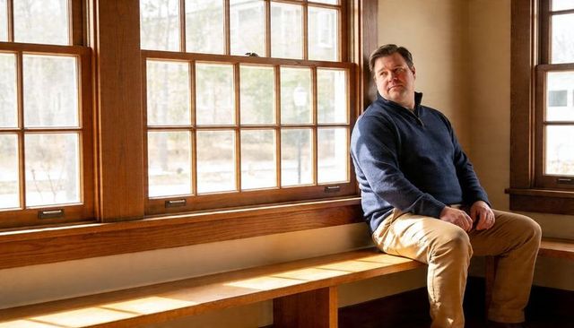 Middle-aged man sitting on window bench gazing out through paned windows, sunlit solitude