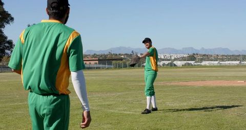 Baseball teammates practicing on sunlit field with mountain backdrop and green uniforms