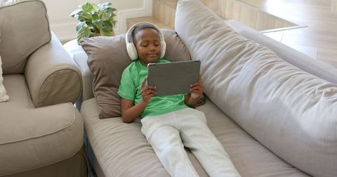 African American boy relaxing on sofa using tablet and headphones for digital learning