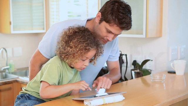 Father and son enjoying a touchscreen tablet in a modern kitchen. Father guiding son with device, making learning fun. Perfect for parenting blogs, technology in family life articles, educational websites, or advertising for family-related products.