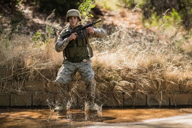 Military soldier in camouflage uniform jumping in water during boot camp training. Ideal for use in articles or advertisements related to military training, physical fitness, tactical exercises, or armed forces recruitment.