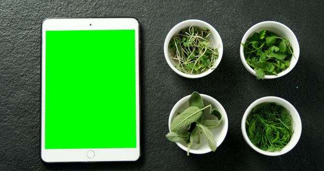 Shows digital tablet with green screen next to fresh herbs in white bowls on dark surface. Tablet screen ready for mockup or advertisement. Ideal for topics on technology in the kitchen, cooking apps, digital recipes, healthy eating, or herbal usage in food.