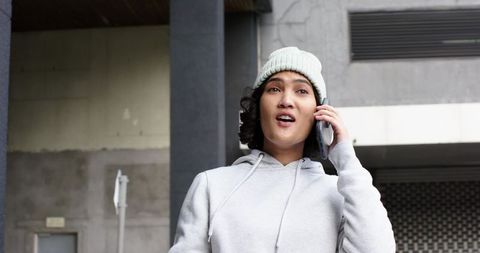 Young woman talking on smartphone outside urban building in hoodie and knit beanie