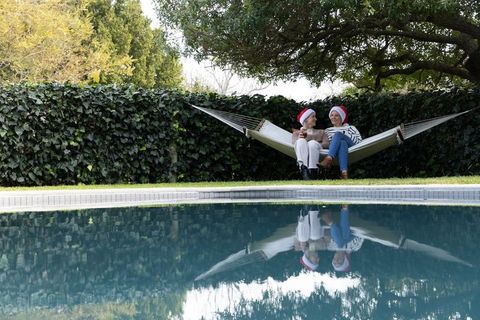 Senior women wearing santa hats sitting on hammock by pool enjoying holiday relaxation