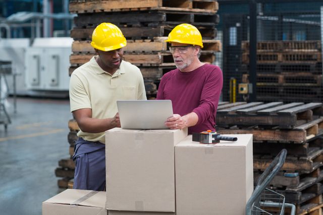 Two factory workers wearing hard hats are collaborating on a laptop in an industrial production plant. They are surrounded by boxes and pallets, indicating a busy work environment. This image can be used to depict teamwork, industrial work settings, logistics, and the integration of technology in manufacturing processes.