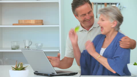 Smiling Senior Couple Online Shopping in Kitchen with Laptop