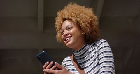 Smiling young woman holding smartphone, looking up in underpass with concrete beams, daylight, strip