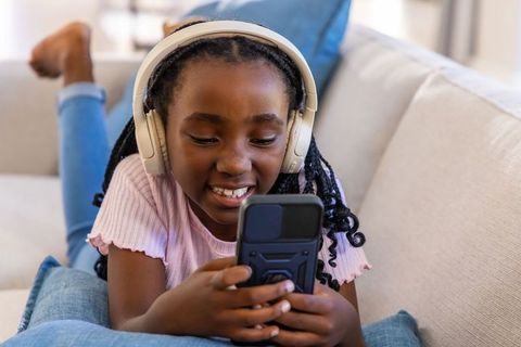 Young african american girl listening on headphones while using smartphone on couch