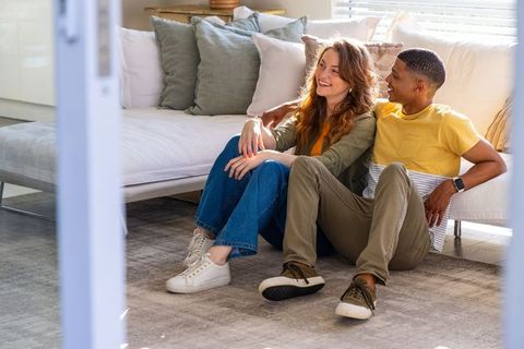 Relaxed interracial couple sitting on rug smiling beside white sofa in bright living room