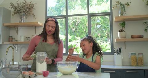 Mother and child baking on sunlit kitchen island, whisking batter and pouring flour
