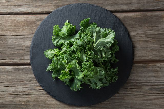 Fresh kale leaves arranged on a round slate plate placed on a rustic wooden table. Ideal for use in health and nutrition articles, vegan and vegetarian recipe blogs, organic food promotions, and superfood marketing materials.
