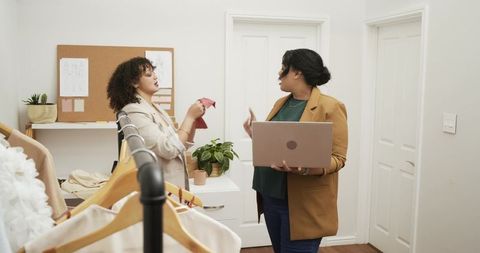 Boutique colleagues reviewing fabric swatch while holding laptop by clothing rack