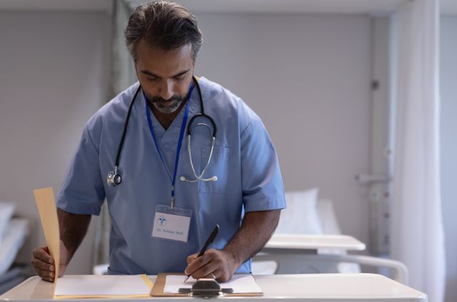 Biracial male doctor in blue medical uniform writing on a clipboard in a hospital ward with a stethoscope around his neck. Suitable for healthcare, medical field promotions, and hospital-related content.