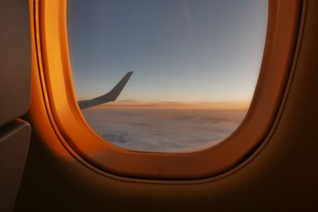 A tranquil view from an airplane window during sunset, showcasing a wingtip set against a blanket of clouds. The sunlight casts a warm glow over the scene. Ideal for themes related to travel, aviation, and serene moments experienced during flights. Perfect for use in travel blogs, airline advertisements, and posters promoting the beauty of flying.