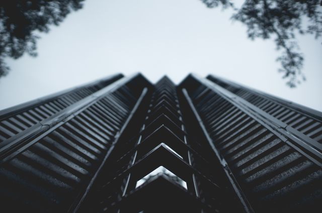 Low angle view showcasing a modern skyscraper silhouetted against an overcast sky, creating a dramatic effect highlighting the geometric lines and structure of the building. Ideal for use in architectural presentations, real estate promotion, urban development advertisements, and articles focusing on city infrastructure.