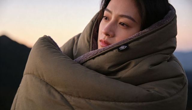 Gazing woman wrapped in olive puffer blanket on mountain ridge at sunrise, cozy outdoor portrait