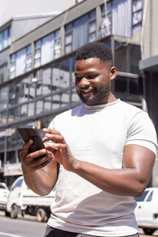 African man smiling while using smartphone on sunny urban street with glass facades