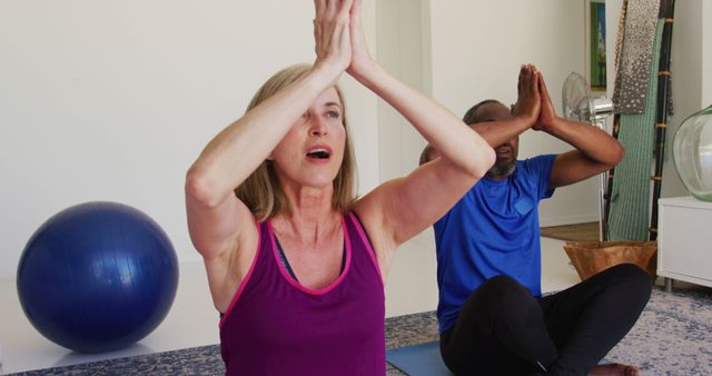 This image depicts a diverse group of adults practicing yoga and meditation indoors. The focus is on a woman wearing a purple tank top in the foreground, with a man in a blue shirt in the background, participating in a yoga meditative pose. Ideal for promoting wellness classes, fitness centers, home workout routines, and mindfulness programs.