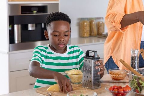 African american child grating cheese with caregiver at modern kitchen island