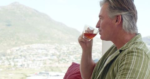 Middle-aged man sipping whiskey by window overlooking mountain town, contemplative lifestyle moment