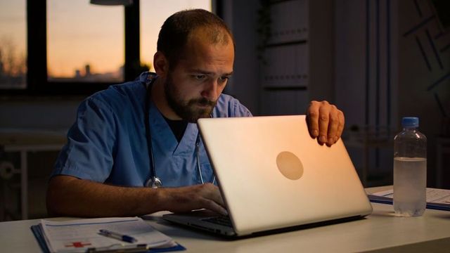 Clinician in scrubs reviewing patient records on laptop at dusk during late shift