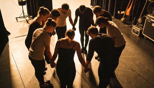 Theatre ensemble holding hands in unity circle under warm spotlight before performance