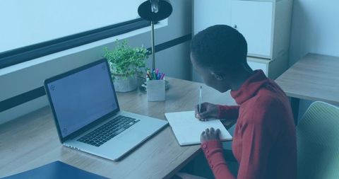 Young woman writing at minimalist home desk with laptop and notebook, focusing on study