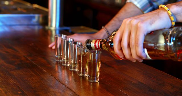 Bartender pouring shots of liquor into clear glasses lined up on a wooden bar counter. Ideal for articles or ads related to nightlife, bar culture, social outings, bartending services, and alcoholic beverages.