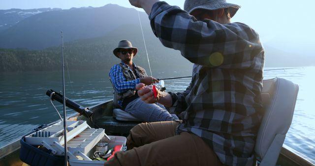 Two middle-aged Caucasian men are enjoying a fishing trip on a serene lake, with copy space. One is preparing a fishing line while the other sits back, both surrounded by tranquil waters and mountain scenery, creating a peaceful outdoor experience.