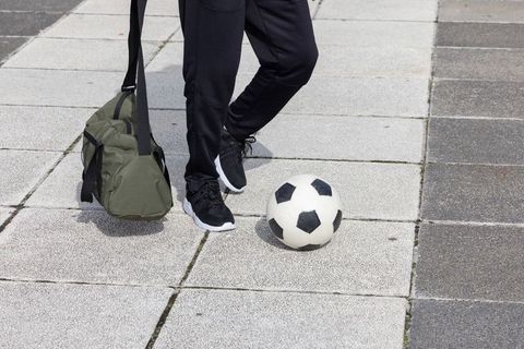 Standing athlete with soccer ball and duffel bag on urban concrete tiles
