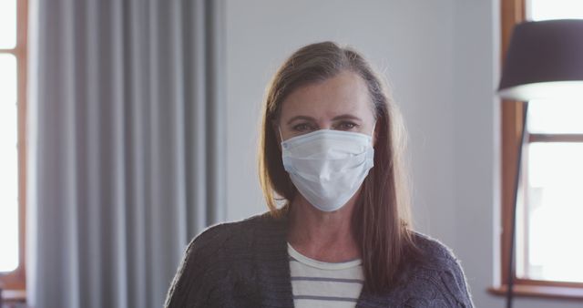 Mature woman standing indoors wearing a surgical mask, illustrating themes of pandemic safety, health protection, and preventative measures during COVID-19. Image useful for articles on public safety, health guidelines, or blogs discussing the pandemic. Suitable for medical advertisements, educational materials, and public health announcements.